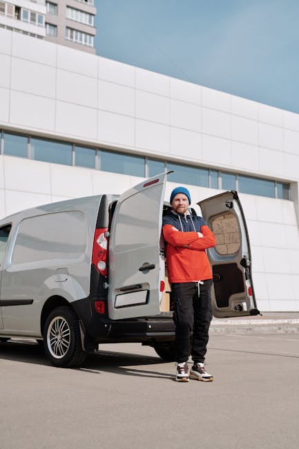 A man dressed in a red and navy jacket, black trousers, and a blue beanie hat stands with arms crossed next to a grey cargo van with open rear doors in a modern urban setting during daytime. The back 