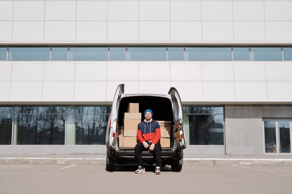 Close-up view of a man with a beard and wearing a black beanie and dark hoodie with red accents, sitting inside a white van during a home relocation process. The man is positioned behind the steering 