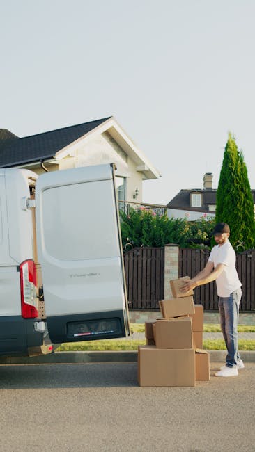 A male mover sits on the edge of an open van door during a home relocation in Tufnell Park. He is dressed in a blue beanie, a navy and red waterproof jacket, black trousers, and white sneakers with bl